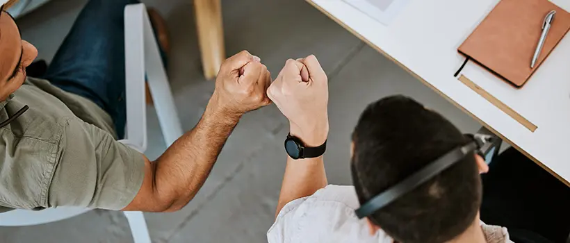 A photo looking down at two employees bumping fists while sitting at a table.