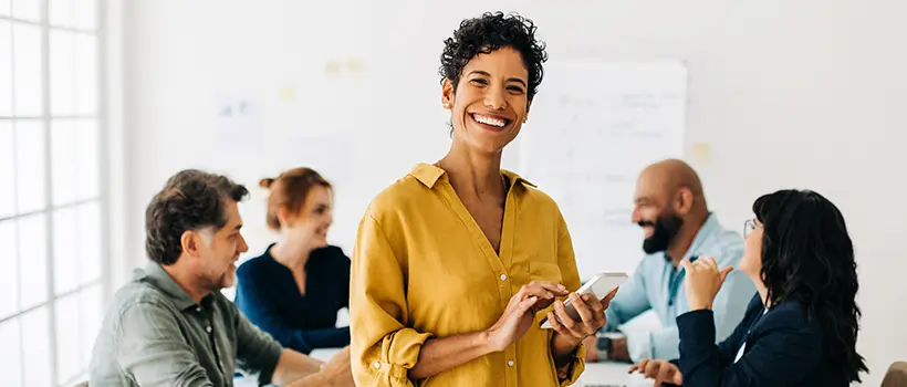 A photo of a woman smiling while holding a phone in front of her employees who are speaking together at a conference table.