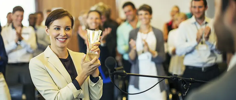 A photo of a businesswoman giving an award to an employee in front of colleagues.