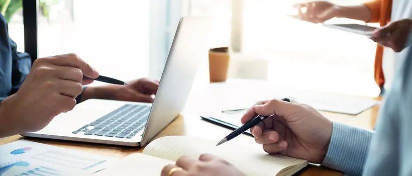 A photo of a person reviewing a notebook while another individual works on a laptop. 