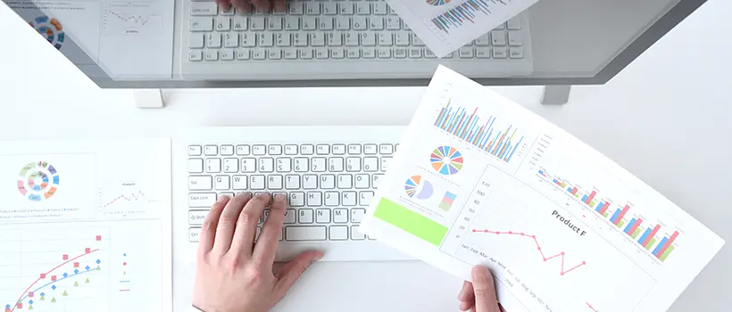 A photo of a person looking at a paper with graphs while typing on a laptop.