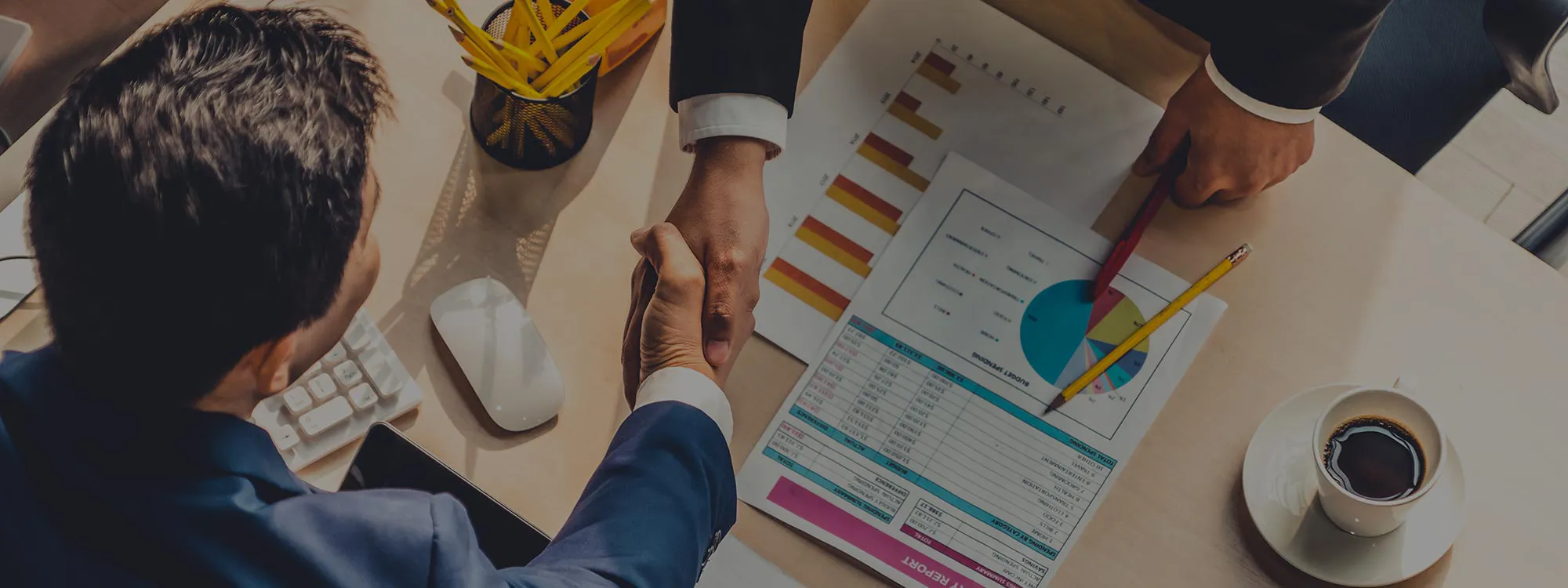 An overhead photo of 2 individuals shaking hands over a desk covered with reports.