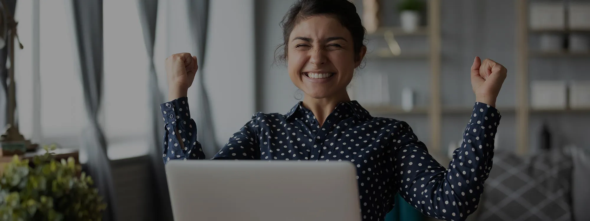 A photo of a woman at a laptop with her hands raised in celebration.